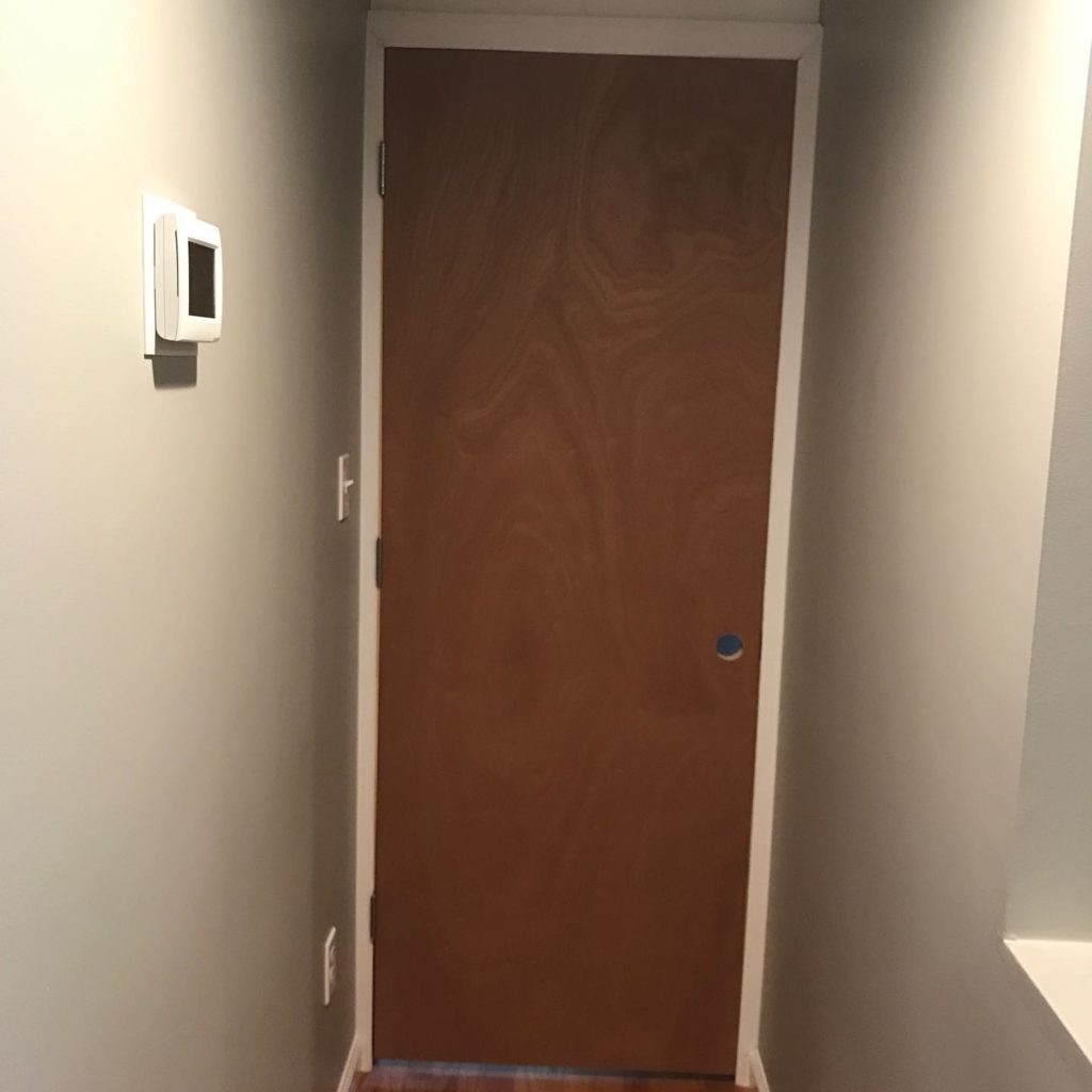 Narrow hallway with a closed wooden door, light gray walls, and a wall-mounted thermostat.